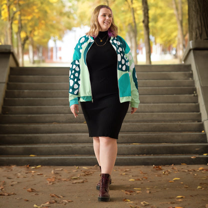 Good Wishes 365 unisex bomber jacket with blue and mint green abstract print worn over a black ribbed dress outdoors.