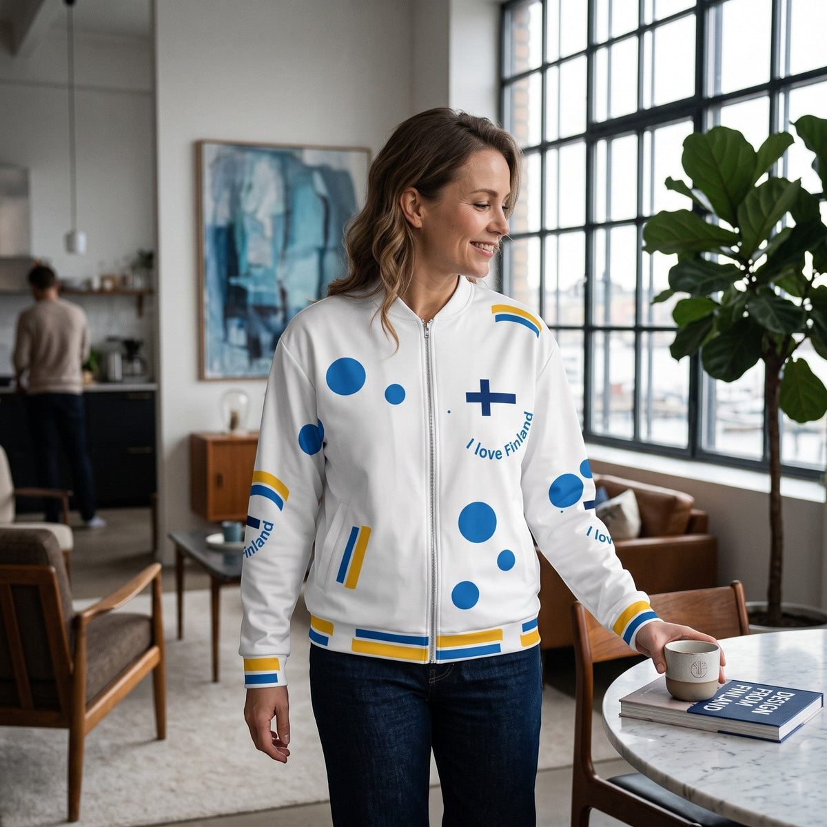 A woman wearing a Good Wishes 365 white bomber jacket with blue and yellow Finland-themed patterns in a bright room.