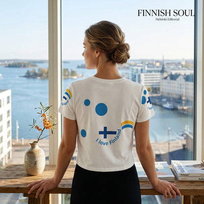 Good Wishes 365 white crop tee with Finnish flag and blue dot graphics, worn by a woman looking out a window.