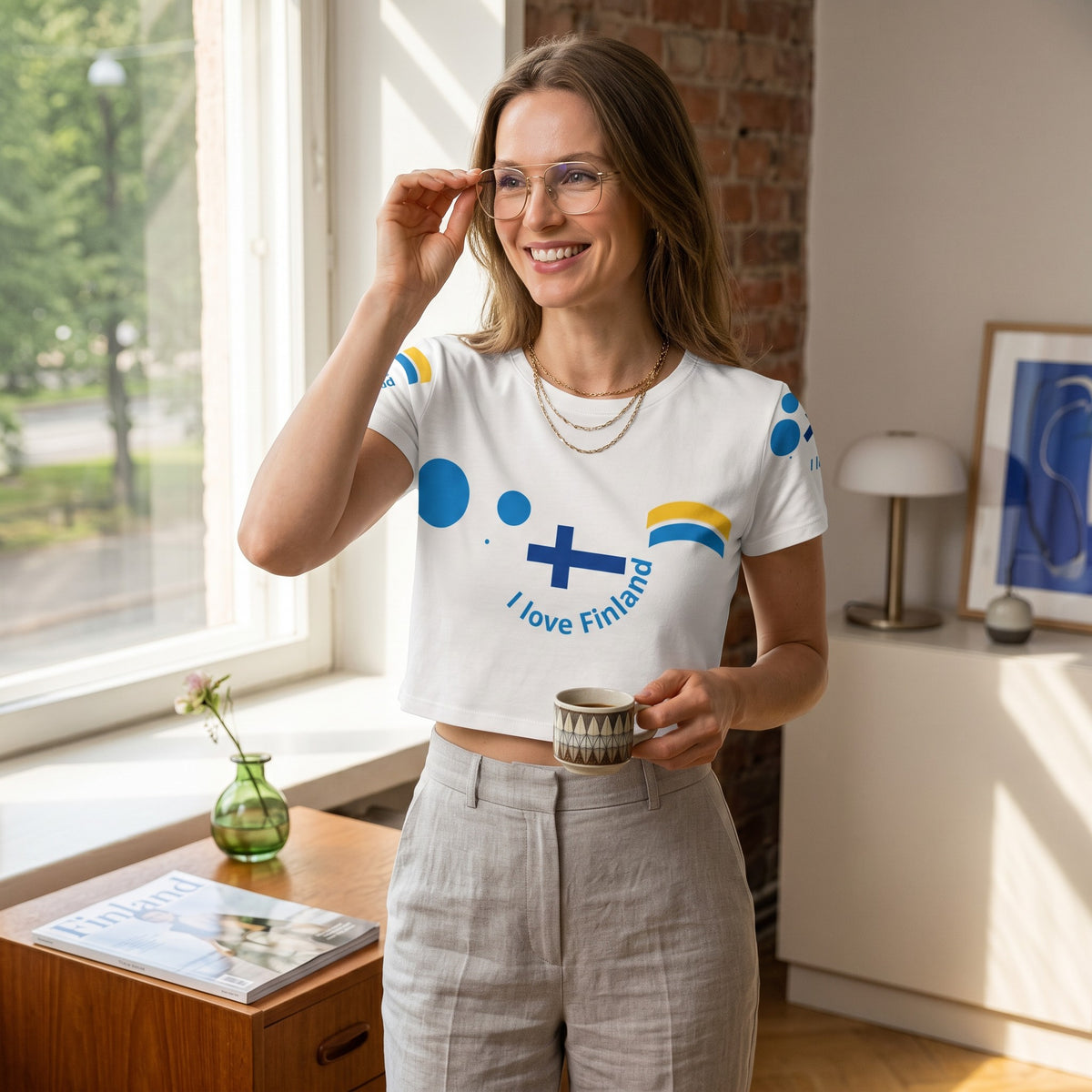 Good Wishes 365 women's white crop tee with Finnish flag design, worn by a smiling woman in a bright room.