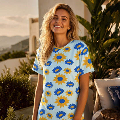 A smiling woman wearing a Good Wishes 365 light blue t-shirt dress with a yellow and blue floral print outdoors.