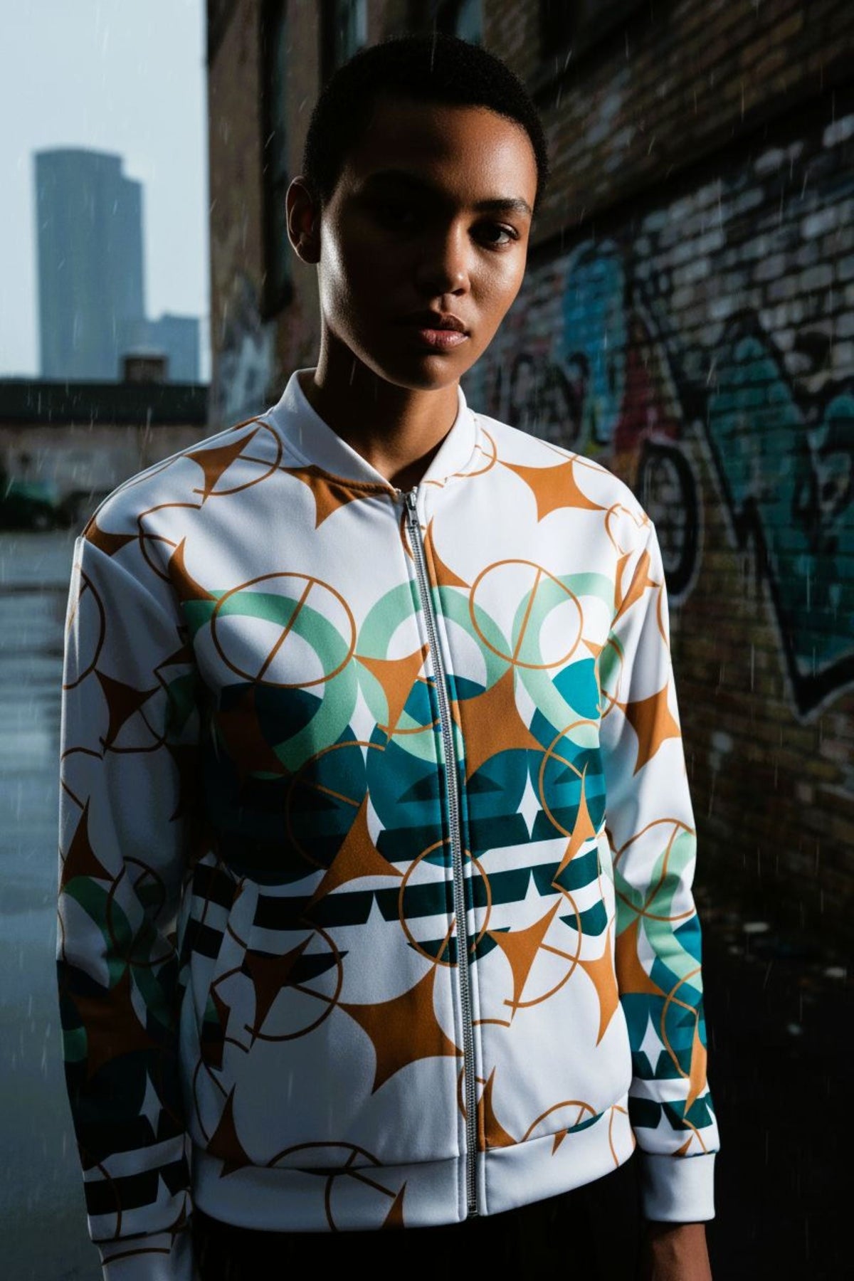 Good Wishes 365 unisex bomber jacket with white base and teal, sage, and ochre geometric star pattern in city setting