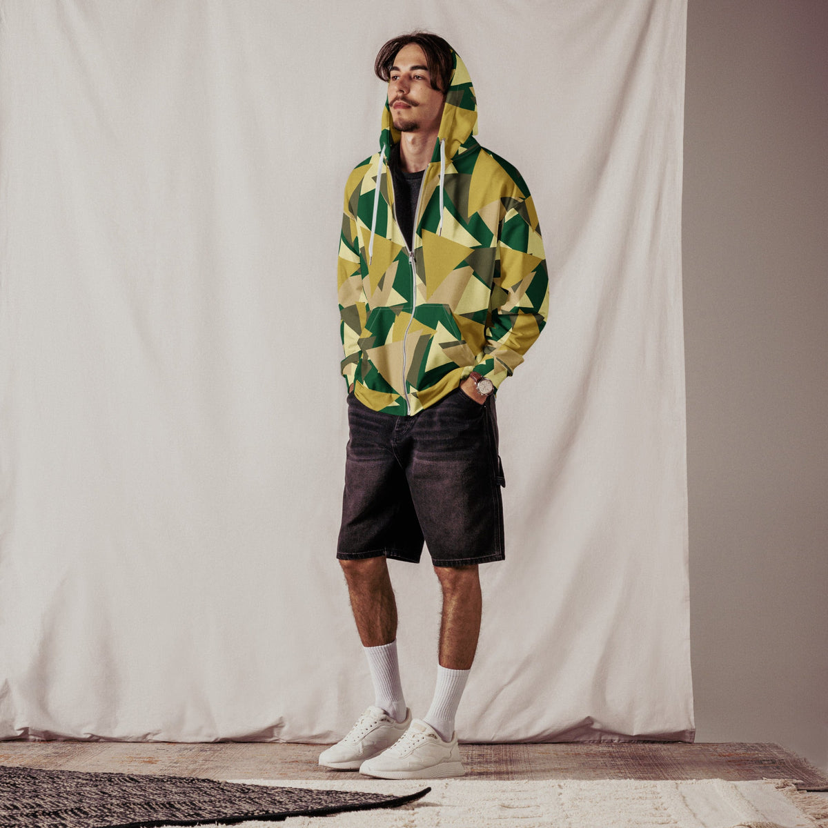 Good Wishes 365 men's zip hoodie with a geometric camo print in green and yellow, worn with black shorts and sneakers.
