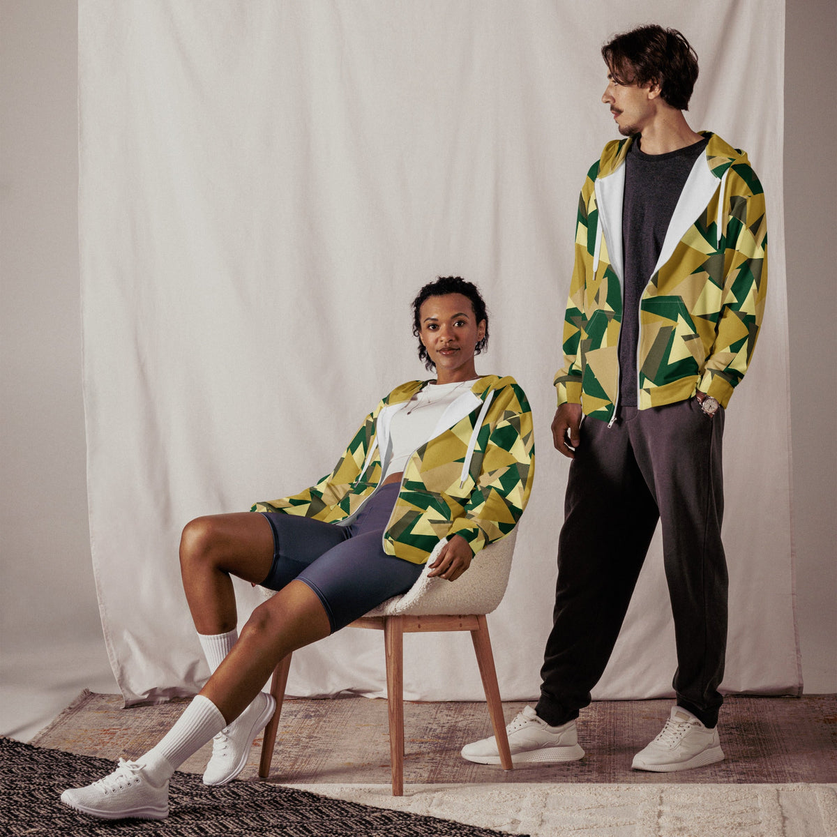 Good Wishes 365 unisex zip hoodie with geometric camo print worn by a man standing and a woman sitting on a chair.