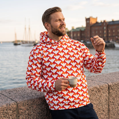 Good Wishes 365 unisex hoodie with red and orange geometric heart pattern, worn by a man holding a coffee by the sea.