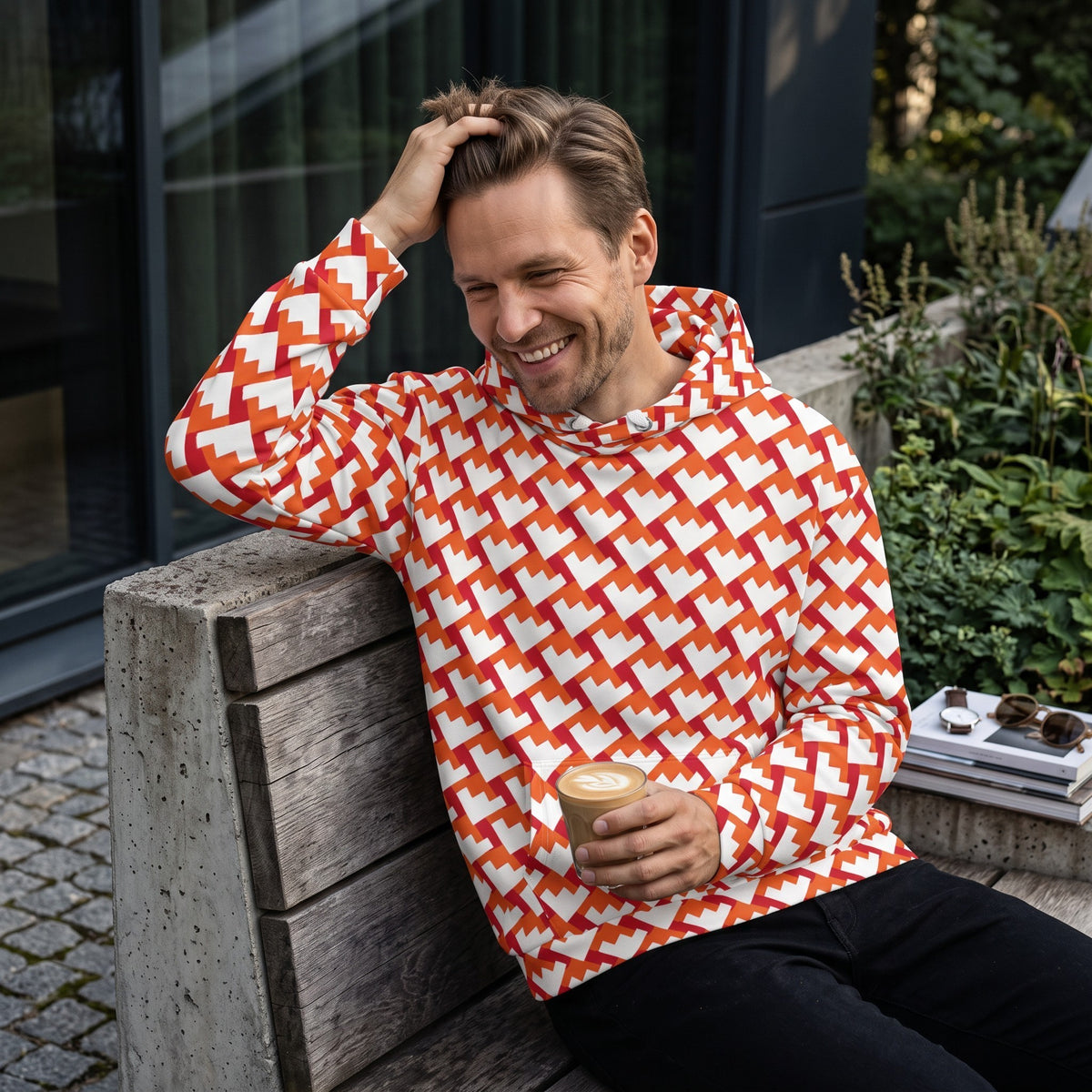 Good Wishes 365 unisex hoodie with a red and orange geometric heart pattern, worn by a smiling man on a bench.