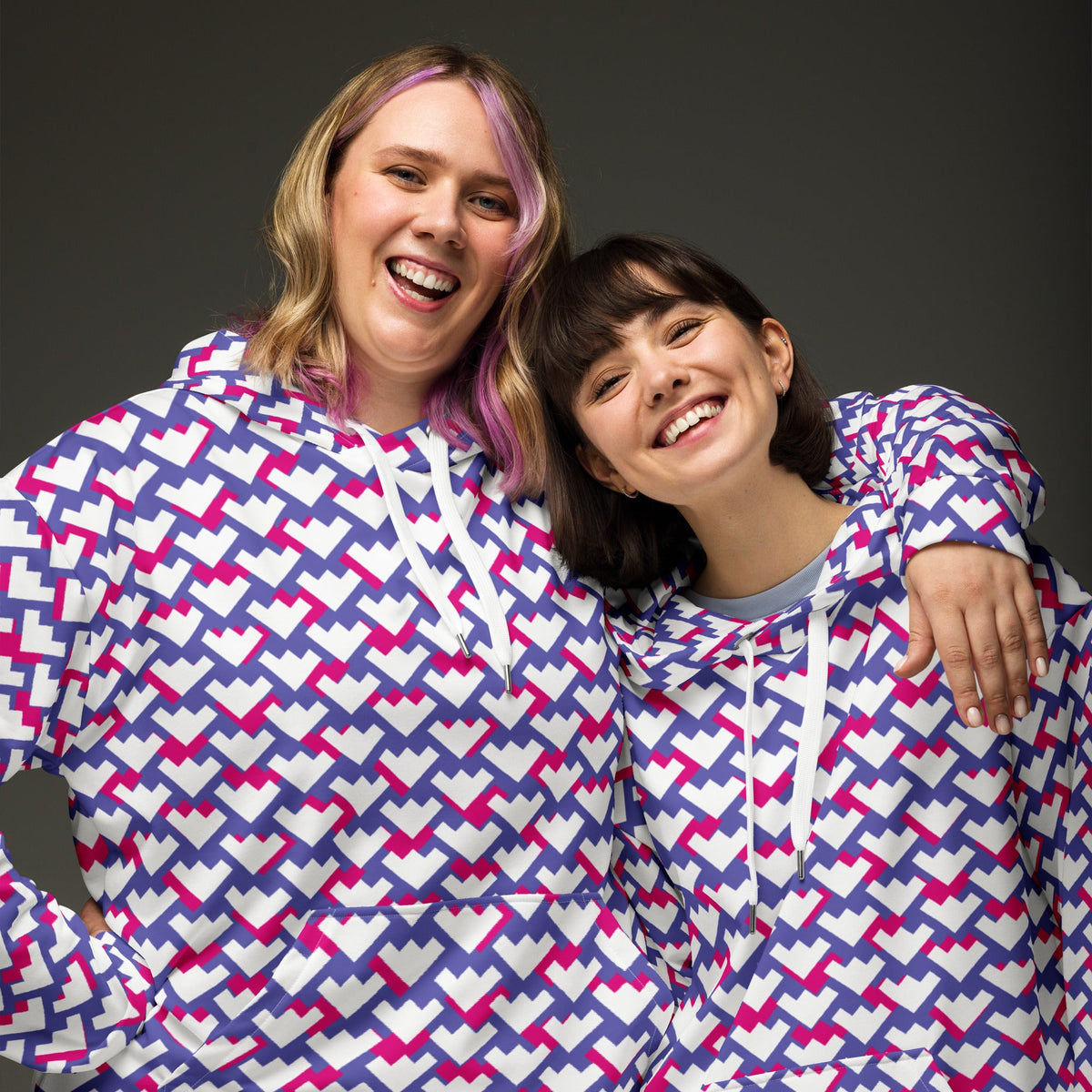 Two smiling women wearing Good Wishes 365 recycled unisex hoodies with a purple and red geometric heart pattern.
