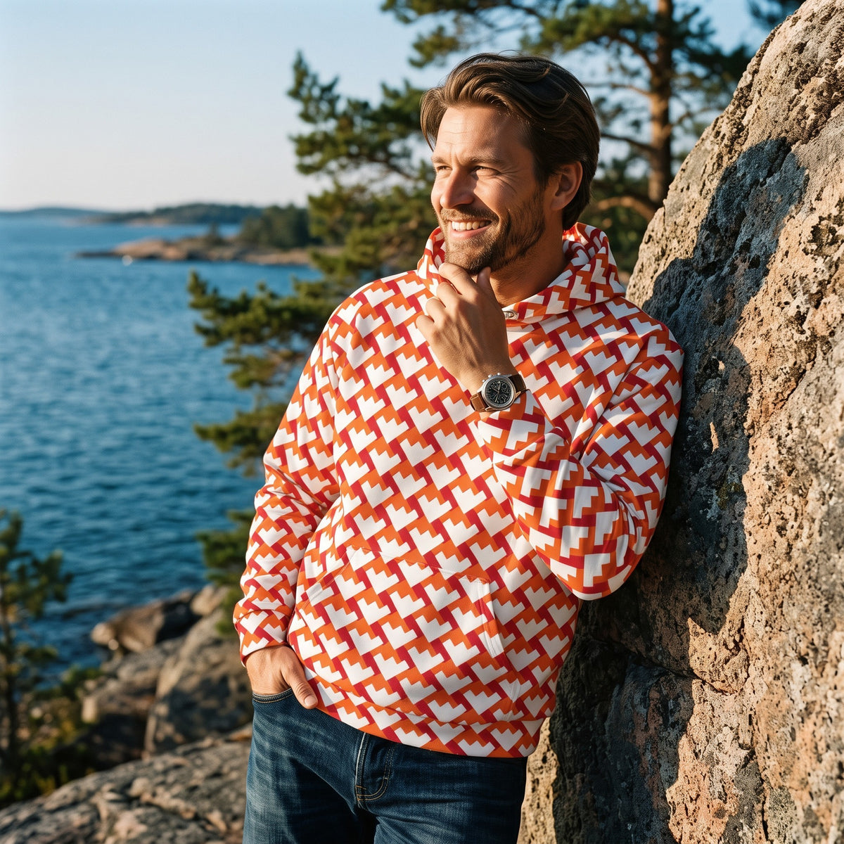 Good Wishes 365 unisex hoodie with red and orange geometric heart pattern, worn by a man leaning on a coastal rock.
