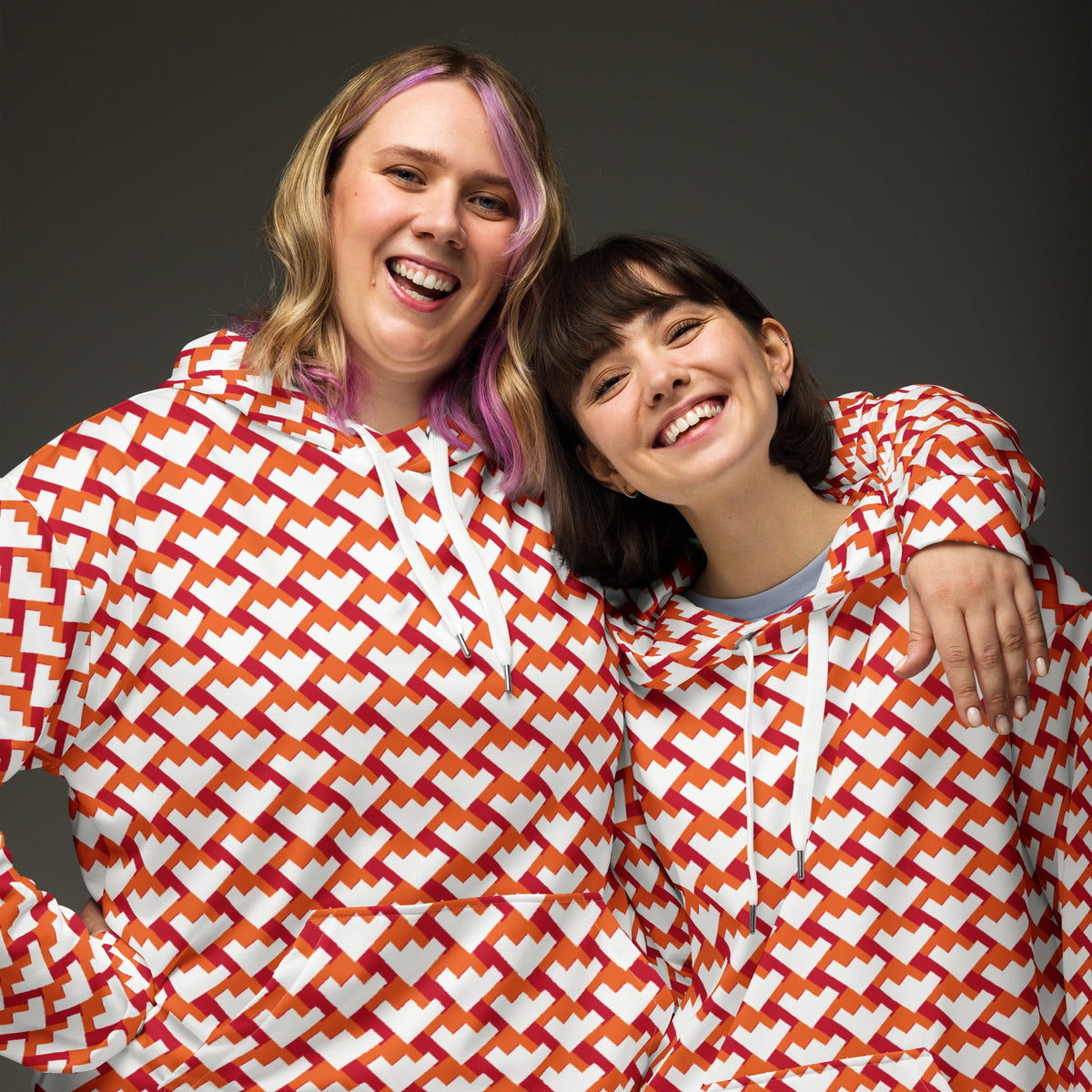 Two women smiling in Good Wishes 365 recycled unisex hoodies with a red and orange geometric heart pattern.