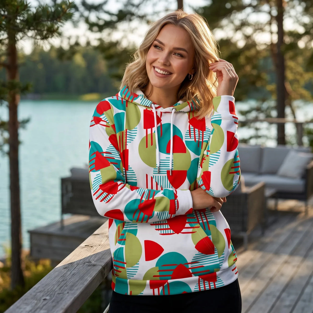 Woman wearing a Good Wishes 365 recycled hoodie with a vibrant red, green, and teal geometric pattern by a lake.