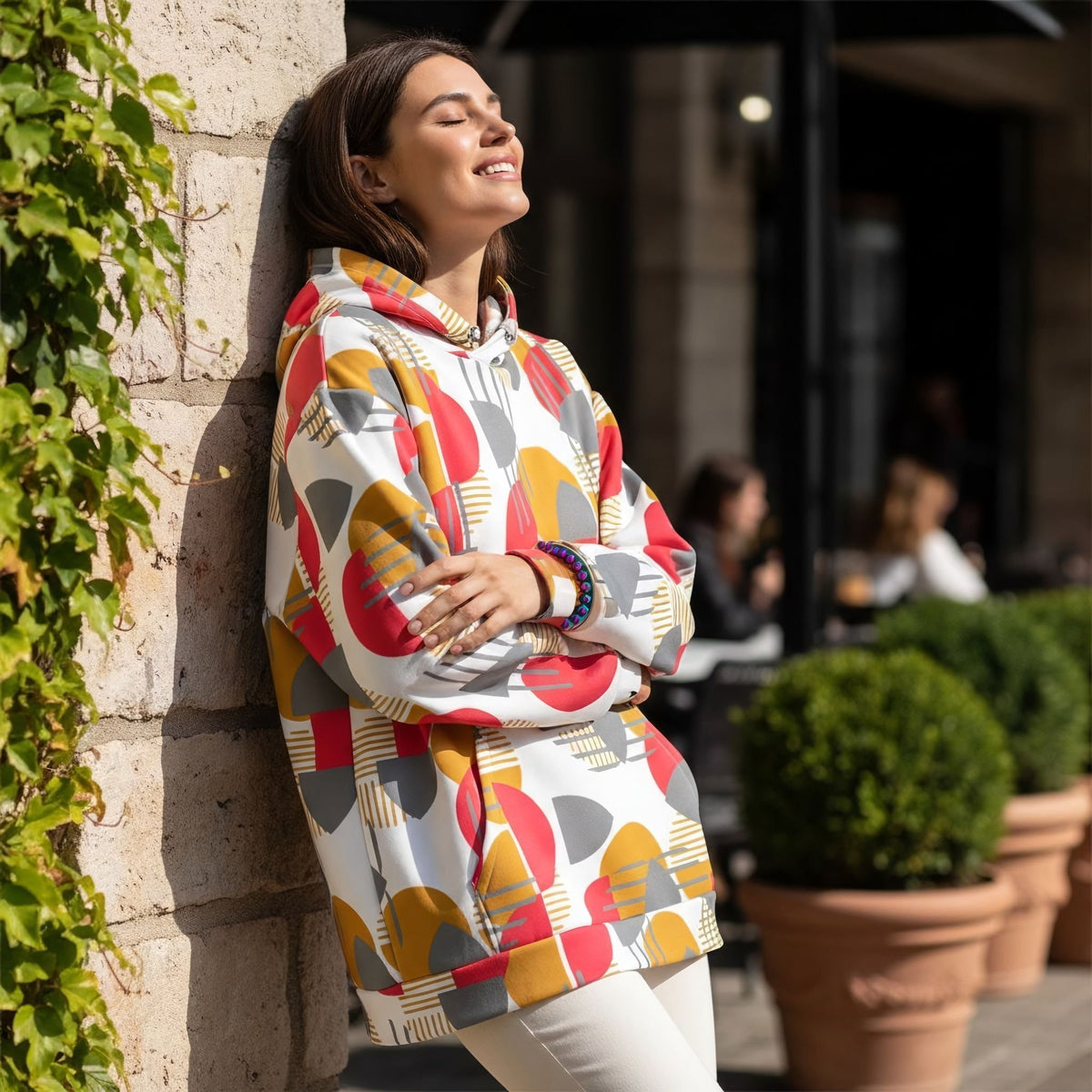 Good Wishes 365 recycled hoodie with rose red, mustard, and silver geometric print worn by a woman leaning on a wall.