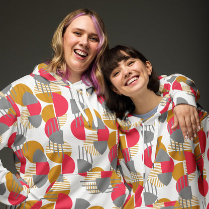 Two smiling women wearing Good Wishes 365 recycled hoodies with a rose red, mustard, and silver geometric print.