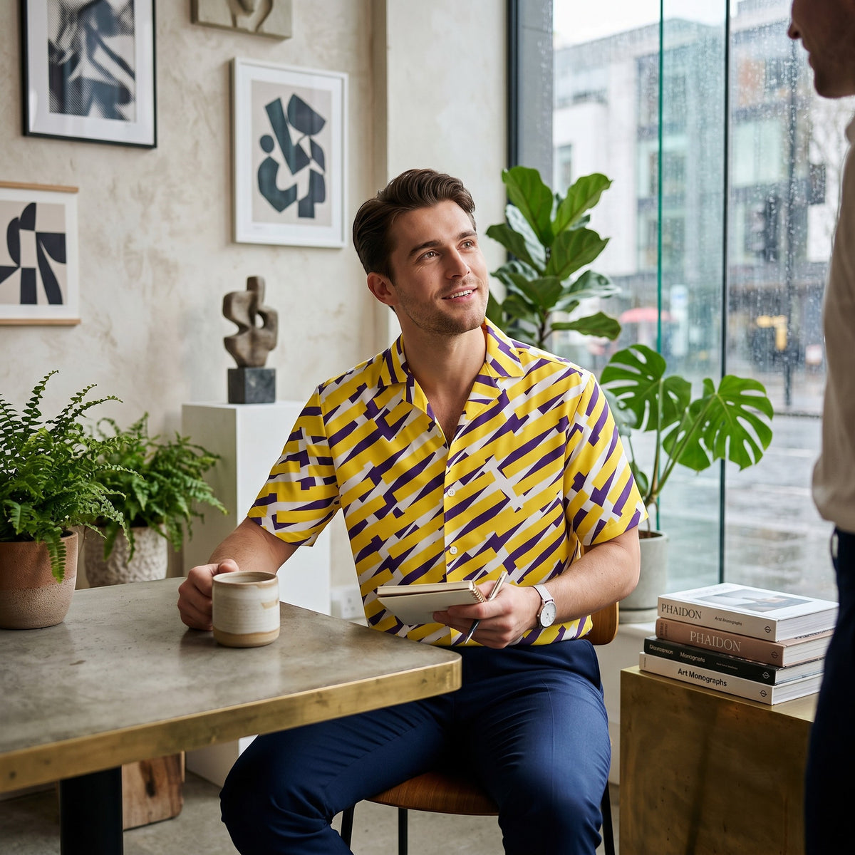 Good Wishes 365 short sleeve button down shirt with yellow and purple geometric print worn by a man in a cafe