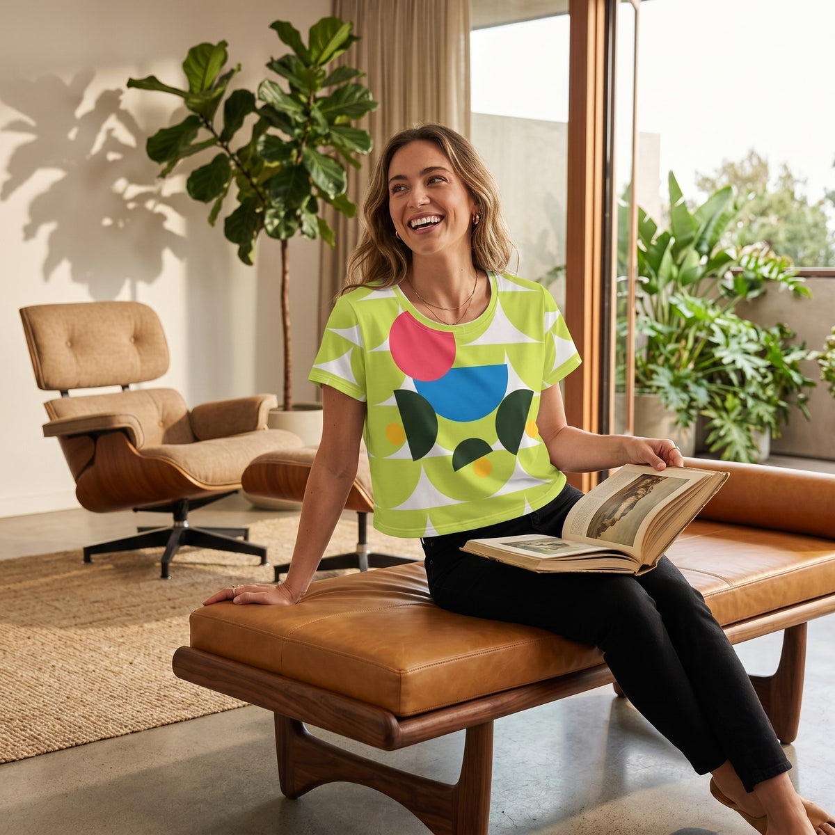 Good Wishes 365 green crop tee with mushroom print worn by a smiling woman sitting on a leather bench in a bright room.