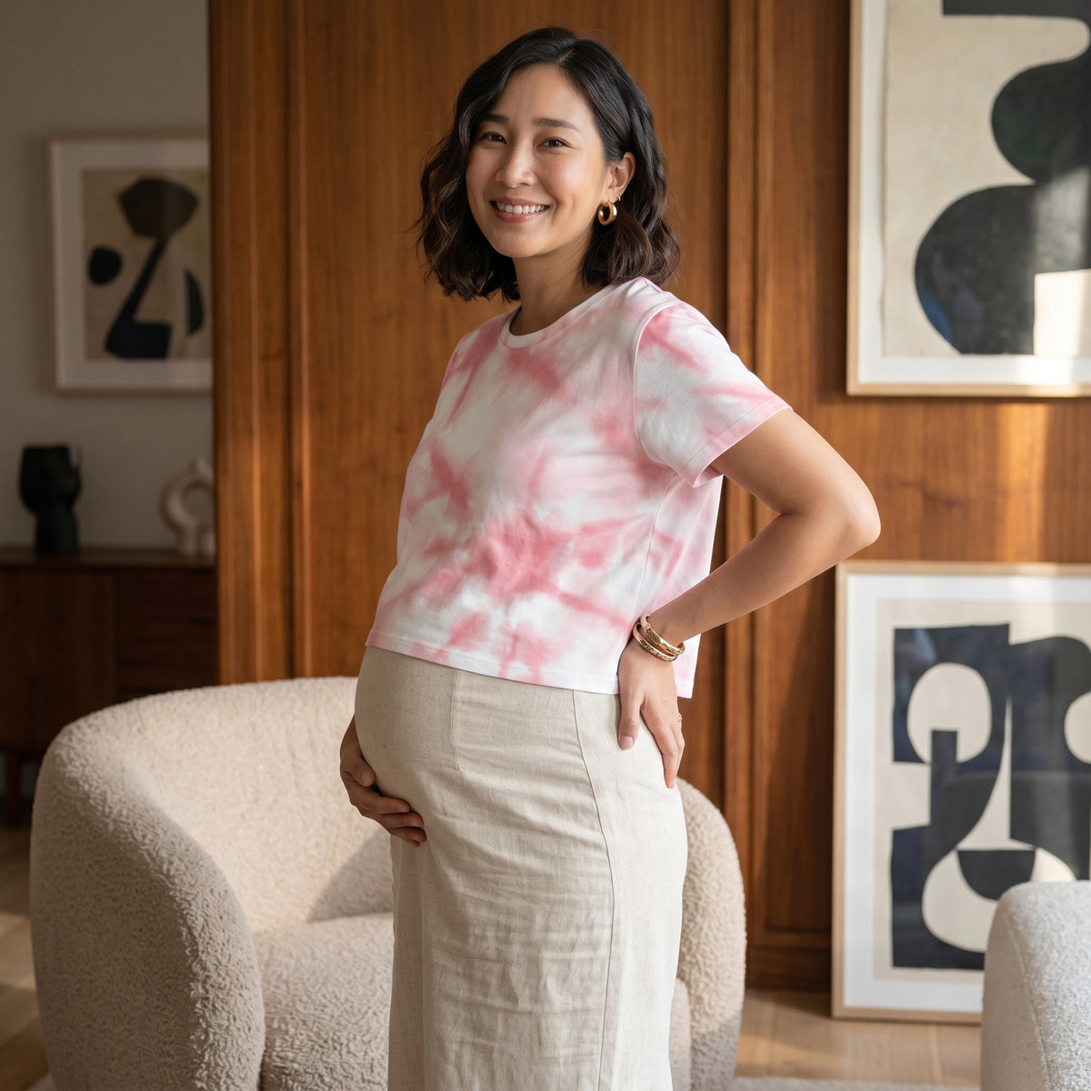 A pregnant woman wearing a Good Wishes 365 pink tie-dye crop tee with a beige skirt in a modern living room.