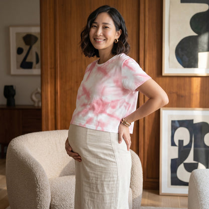 A pregnant woman wearing a Good Wishes 365 pink tie-dye crop tee with a beige skirt in a modern living room.