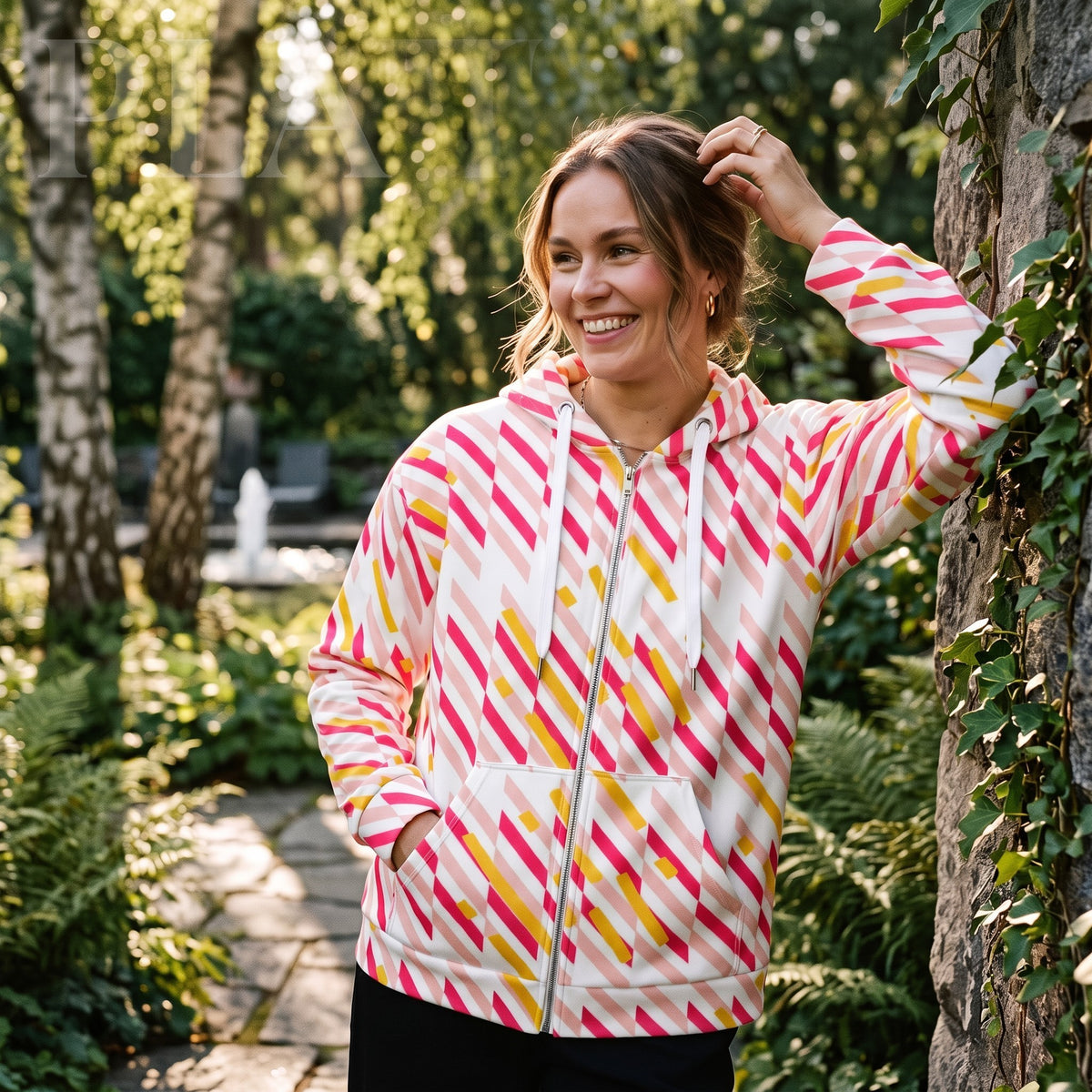 Good Wishes 365 pink and yellow geometric print recycled zip hoodie worn by a smiling woman in a garden setting.
