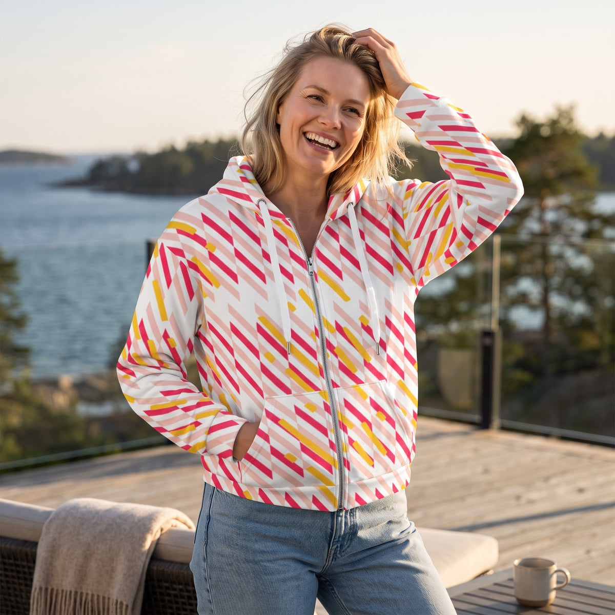 A woman wearing a Good Wishes 365 recycled zip hoodie with a pink and yellow geometric print on a sunny outdoor deck.