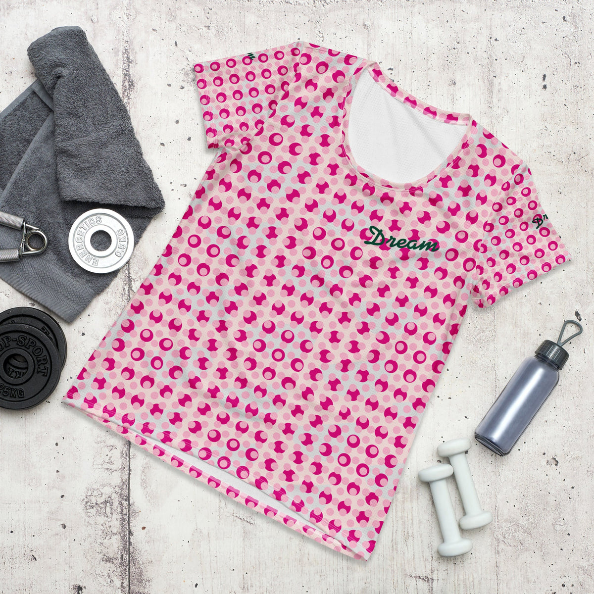 Good Wishes 365 athletic t-shirt with a pink polka dot print and Dream text, laid out with gym gear on concrete.