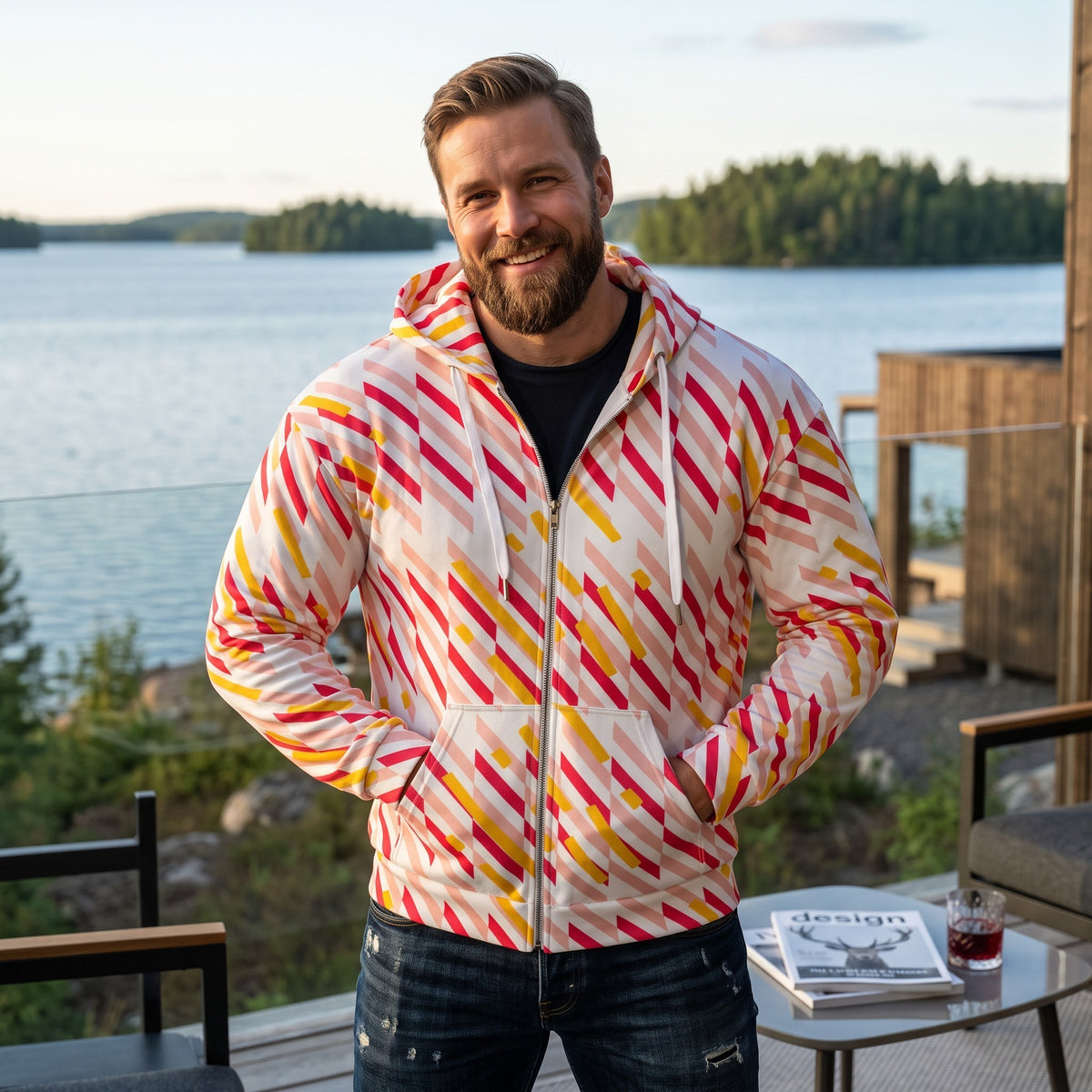 Good Wishes 365 recycled zip hoodie with pink and yellow geometric lines, worn by a smiling man on a lakeside deck.