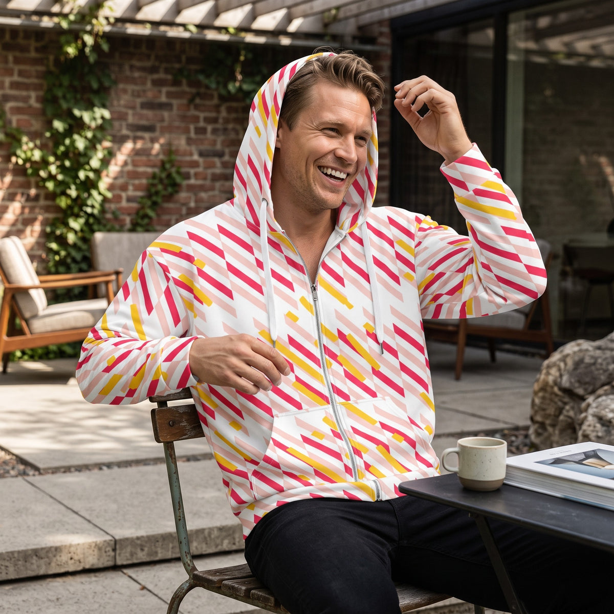 Good Wishes 365 recycled zip hoodie with pink and yellow geometric lines worn by a smiling man on a patio.