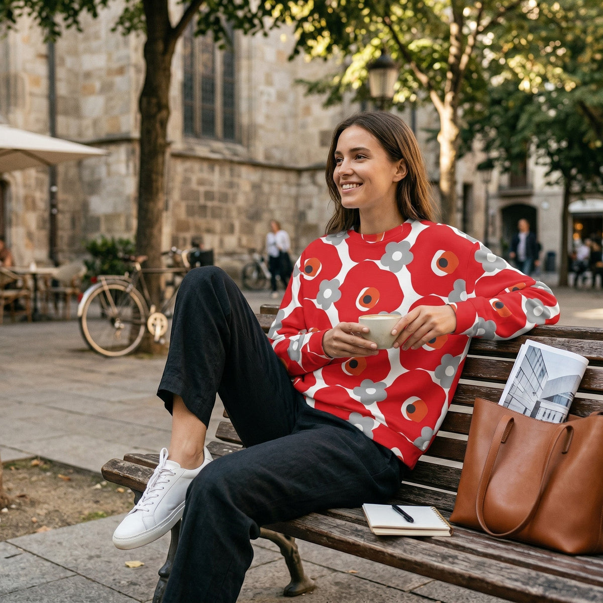 Woman wearing a Good Wishes 365 red and gray floral graphic sweatshirt while sitting on a park bench with coffee.