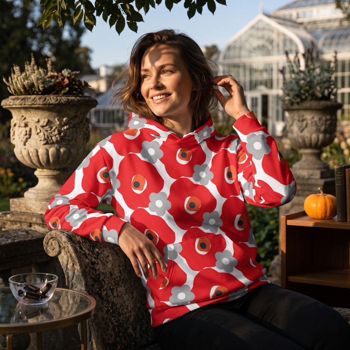 A woman wearing a Good Wishes 365 recycled hoodie with a red and gray floral print in a sunny outdoor garden setting.