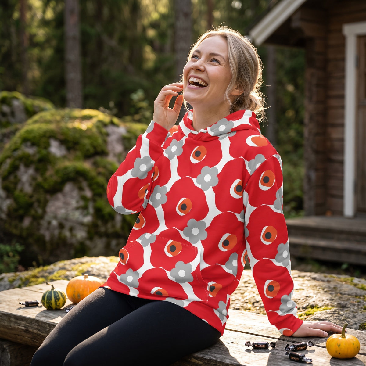 Good Wishes 365 recycled hoodie with red and gray floral print worn by a smiling woman sitting on a wooden bench