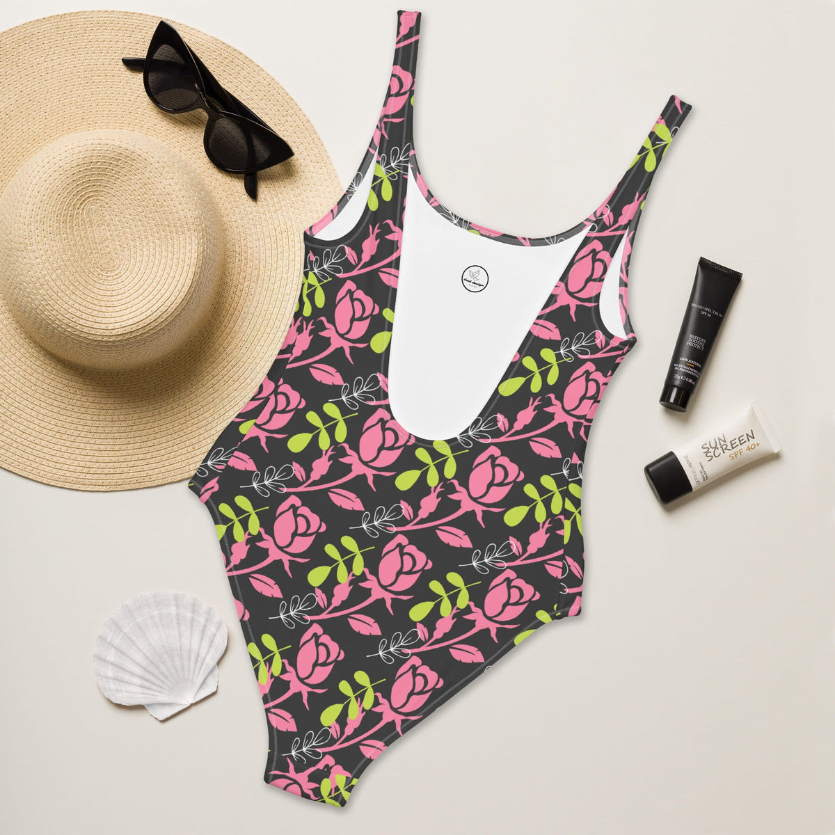 Good Wishes 365 one-piece swimsuit with pink rose print on dark background, styled with straw hat and sunscreen.