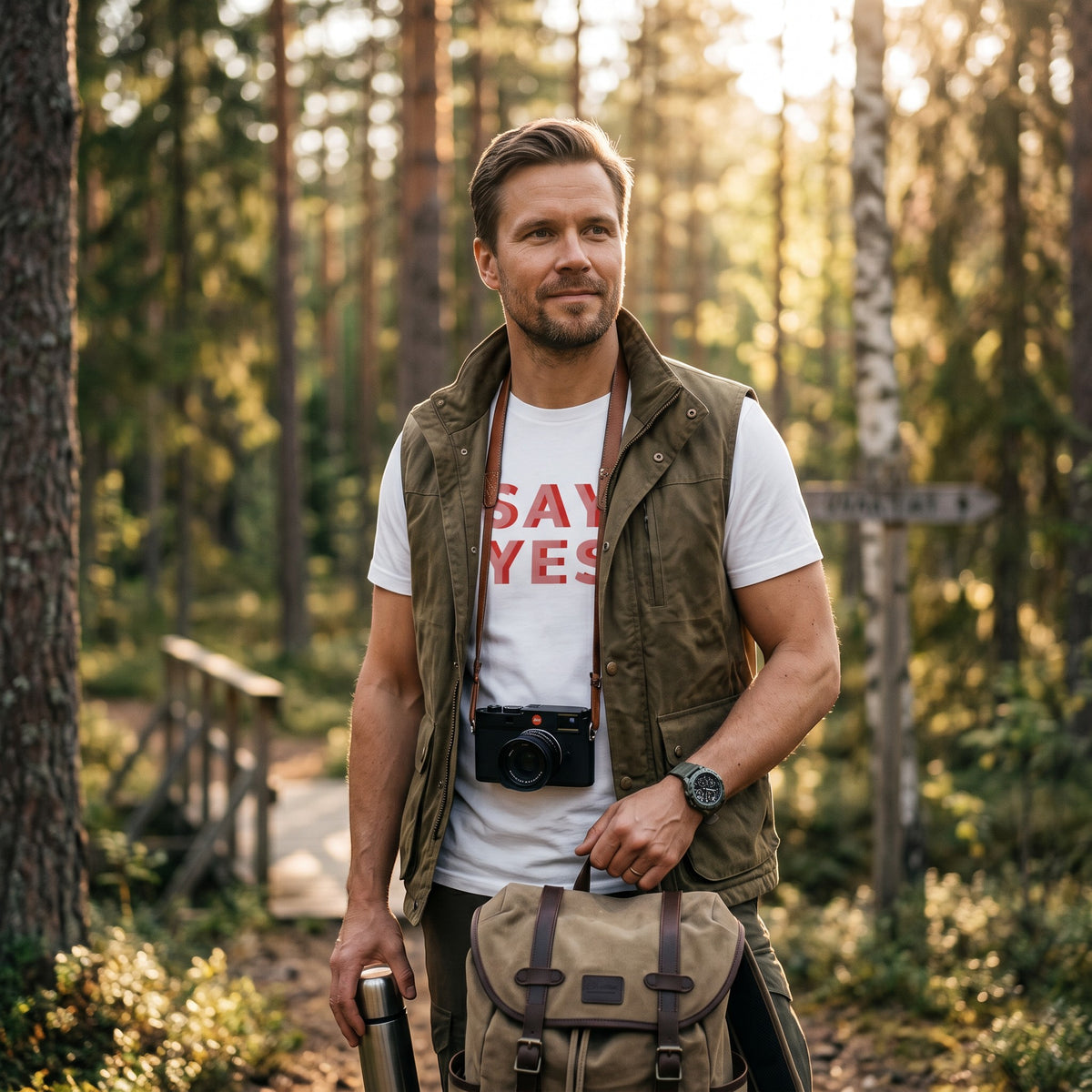Man wearing a white Good Wishes 365 Say Yes graphic cotton t-shirt with an olive vest in a sunlit forest.