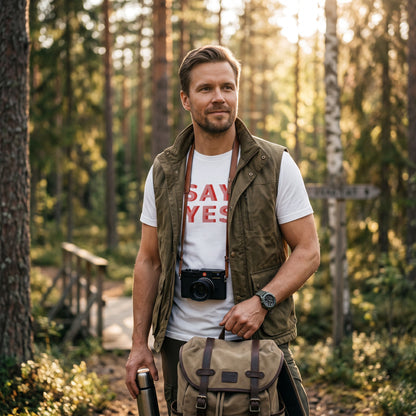 Man wearing a white Good Wishes 365 Say Yes graphic cotton t-shirt with an olive vest in a sunlit forest.