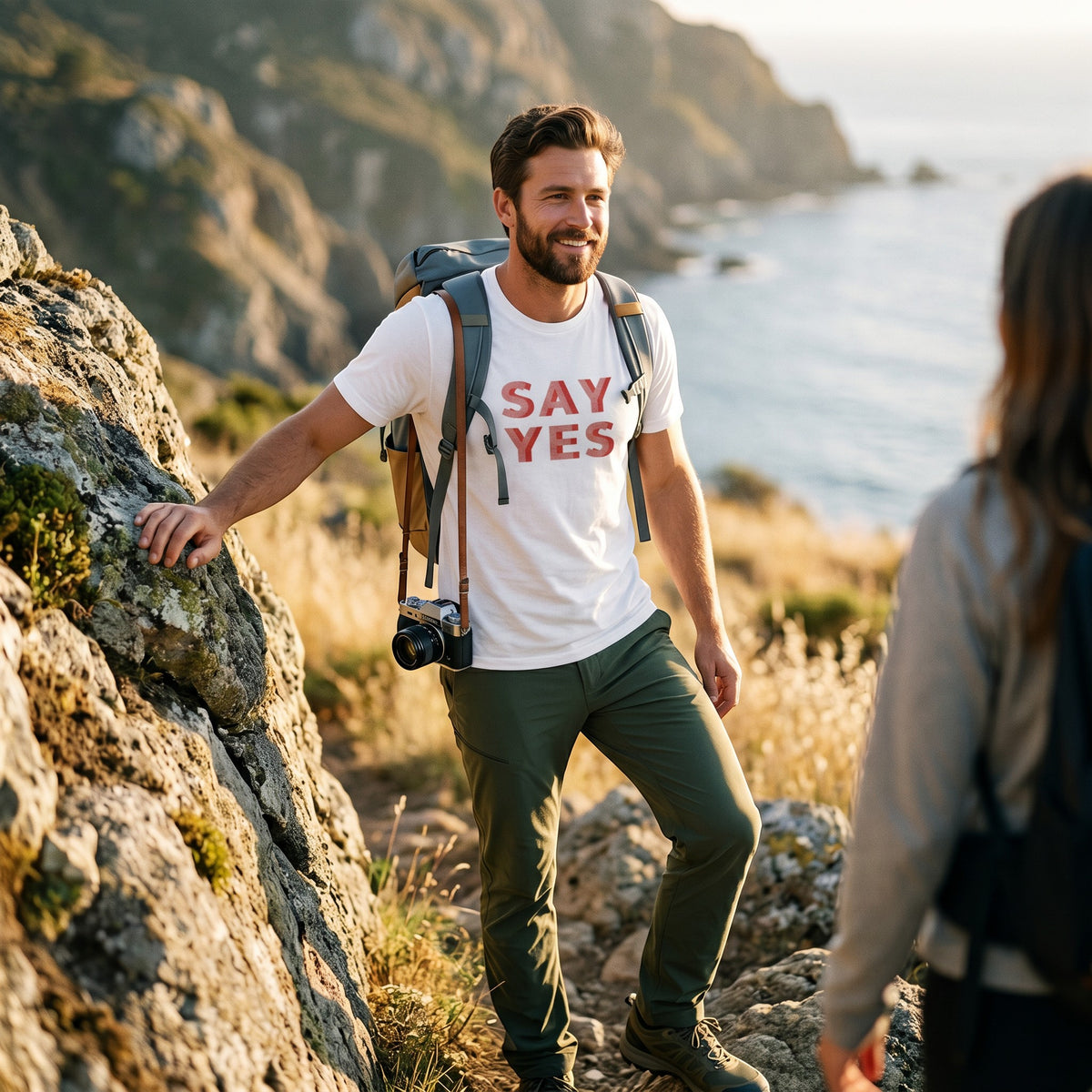 Good Wishes 365 white cotton Say Yes graphic t-shirt worn by a man hiking on a coastal trail with a backpack.