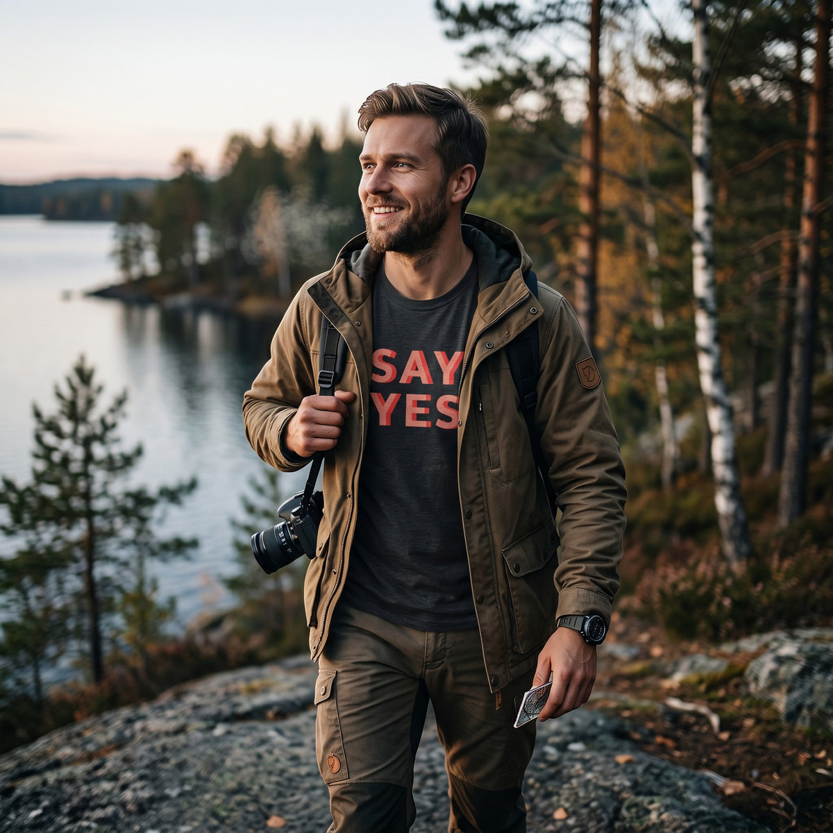 Good Wishes 365 charcoal grey Say Yes graphic t-shirt worn by a man hiking in a forest near a lake.