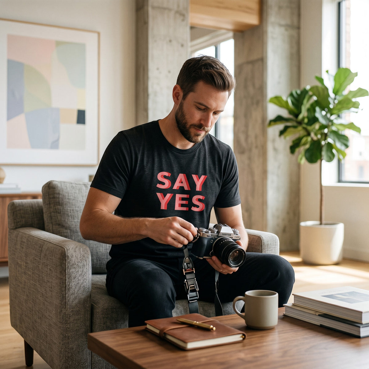 Man wearing a Good Wishes 365 black cotton Say Yes graphic t-shirt while holding a camera in a modern living room.