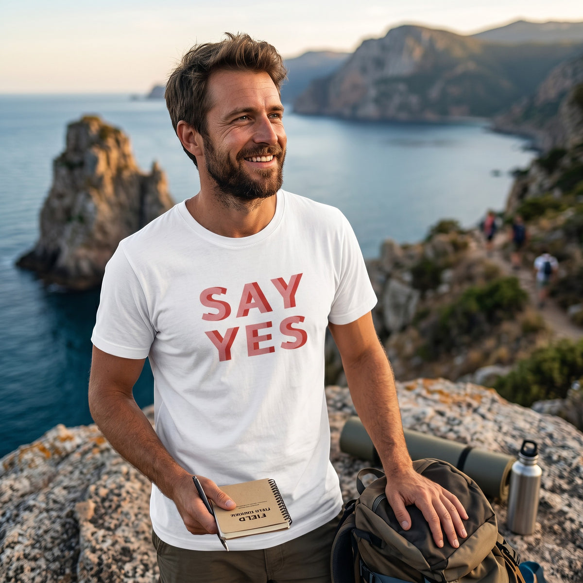 Man wearing a Good Wishes 365 white cotton Say Yes graphic t-shirt while hiking on a coastal cliffside.