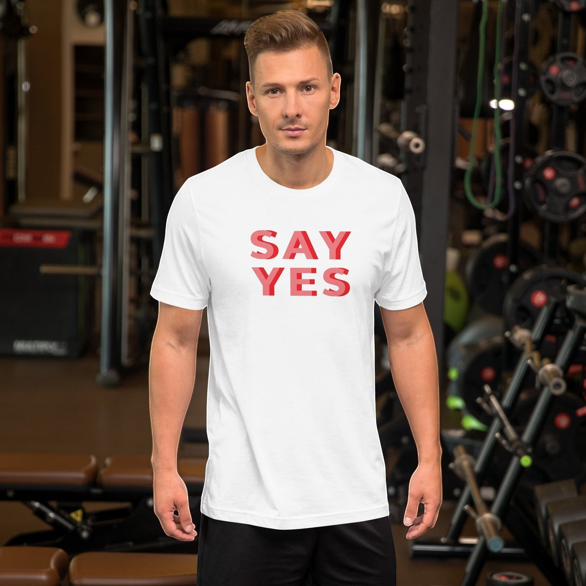 Good Wishes 365 white unisex graphic t-shirt with Say Yes text in coral, worn by a man in a gym setting.