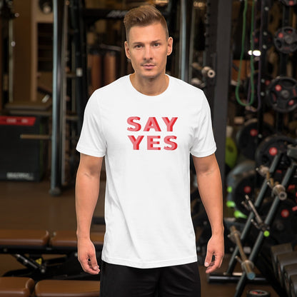 Good Wishes 365 white unisex graphic t-shirt with Say Yes text in coral, worn by a man in a gym setting.