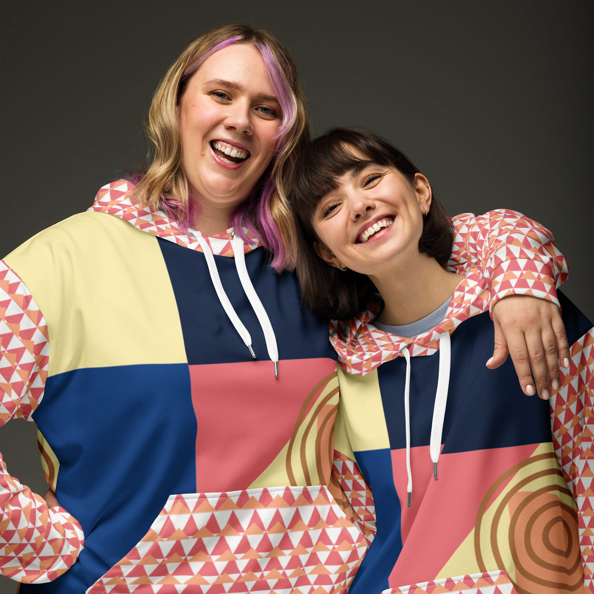 Two women smiling while wearing Good Wishes 365 unisex hoodies with a colorful geometric and abstract print pattern.