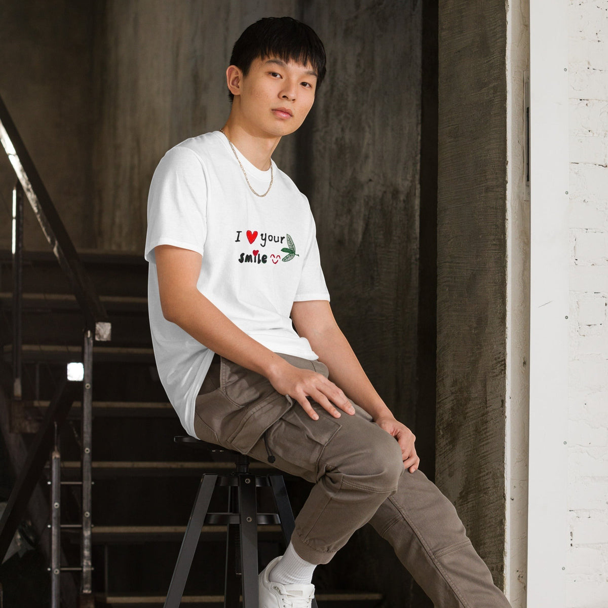 Good Wishes 365 white unisex crewneck t-shirt with I Love Your Smile graphic worn by a man sitting on a stool