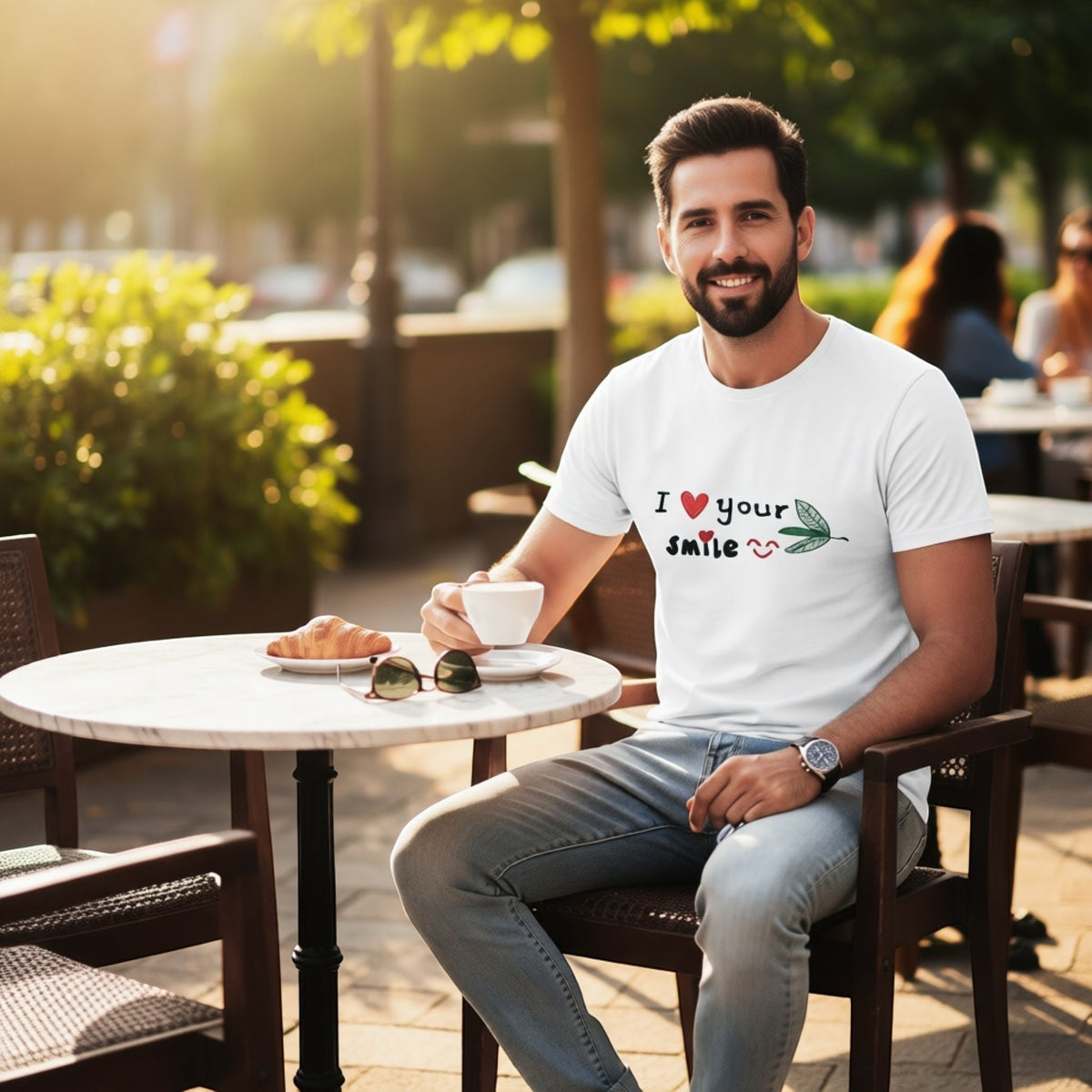 Man wearing a Good Wishes 365 white crewneck t-shirt with I Love Your Smile graphic while sitting at an outdoor cafe.