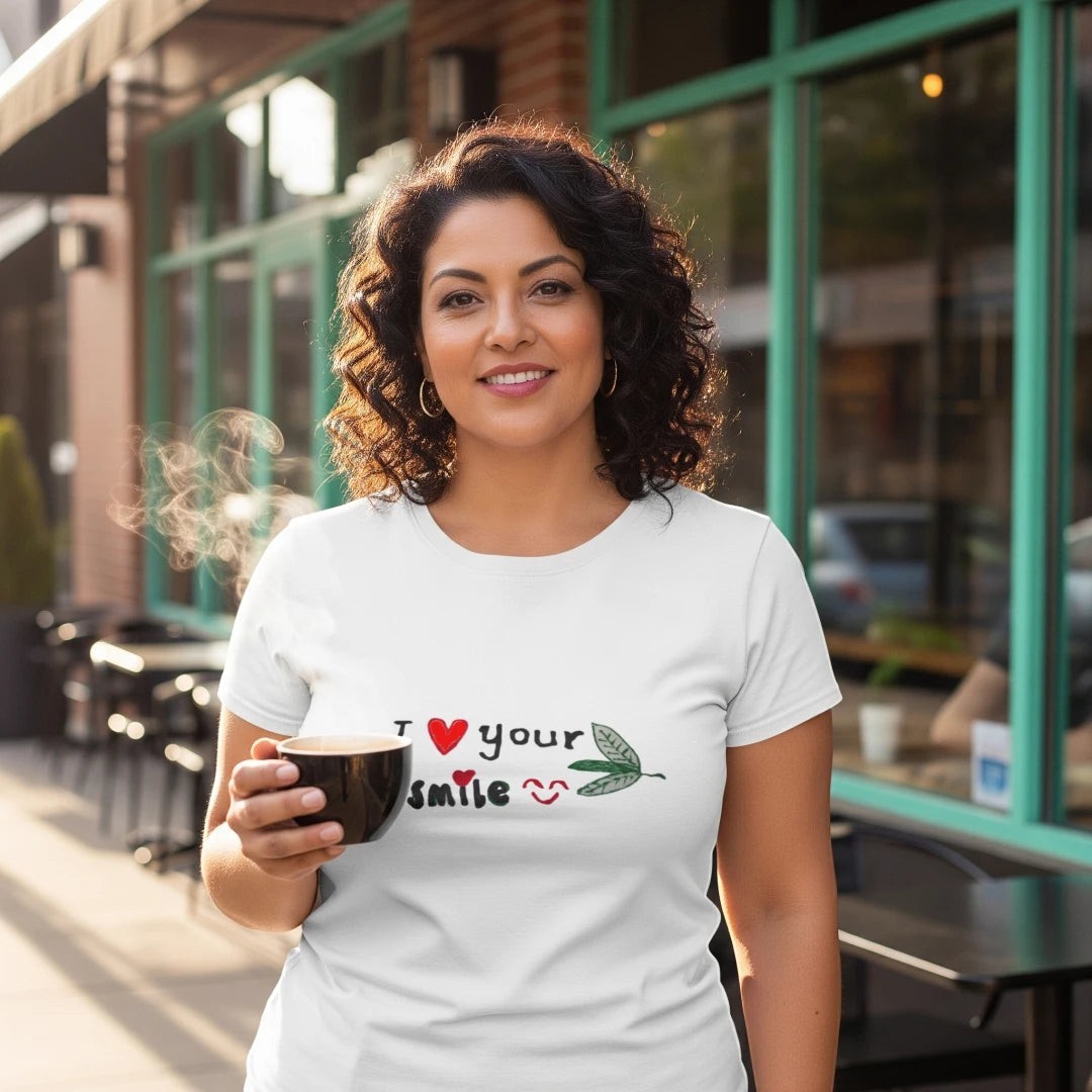 Good Wishes 365 white unisex crewneck t-shirt with I Love Your Smile graphic, worn by a woman holding a coffee cup.